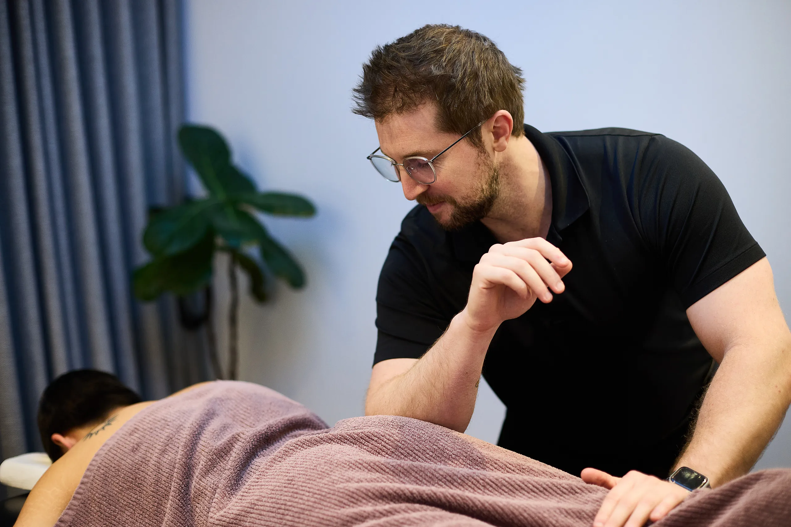 Pure Physio remedial massage therapist Jayke Paulsen prepares to deliver soft-tissue treatment to a client lying face-down beneath a brown towel on the treatment table at the Collins Street clinic, with a fiddle-leaf fig and soft blue curtain in the background.