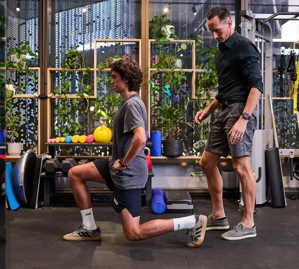 Pure Physio physiotherapist Andrew Sargent guides a young male client through a split-squat lunge during a strength rehabilitation session in the Collins Street clinic gym, surrounded by foam rollers, weighted balls and a plant-lined pegboard wall.