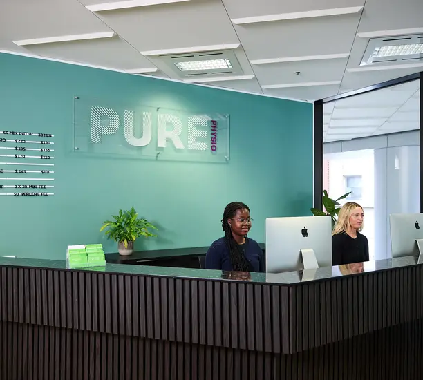 Pure Physio reception and admin team members work at the front desk in the Queen Street clinic, with the turquoise branding wall and green plants visible.
